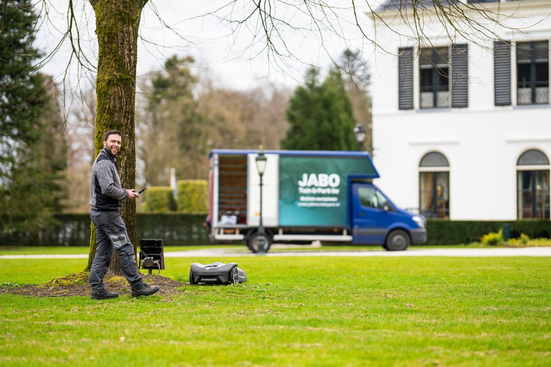 Jaap installeert een robotmaaier bij een landhuis met de JABO-bus op de achtergrond